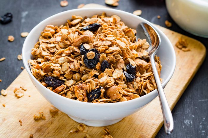 Bowl of granola and dried blueberries with a spoon in it, resting on top of a wooden cutting board.
