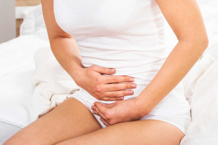 Woman wearing a white tank sitting on her bed clutching her stomach. 